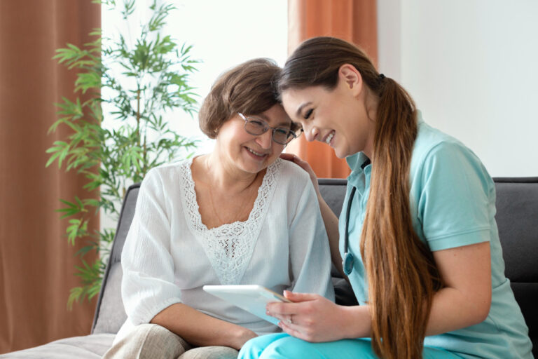 A smiling senior woman sitting on a couch with a caregiver in a blue uniform, both looking at a tablet. The elderly woman is listening about how much does private home care cost per hour and how should she pay for it.